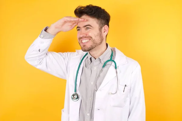 若い白人医師の男性は 孤立した黄色の背景には非常に満足し 遠くに手を頭の上に見て笑みを浮かべて 医療の制服を着て 検索の概念 — ストック写真