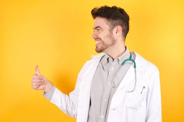 若いハンサムな白人医師の男性は 誇りに思って 側に親指のジェスチャーを行う笑みを浮かべて 医療の制服を着ている いい仕事だ — ストック写真