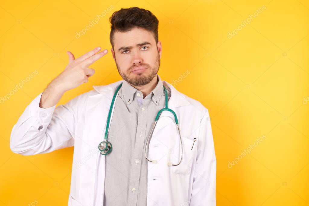Unhappy Caucasian young doctor man imitates gun shoot makes suicide gesture keeps two fingers on temples has puzzled expression dressed in medical uniform stands indoors.