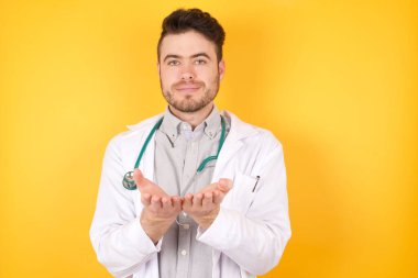 Sarı arka planda poz veren genç beyaz bir doktor avuçlarını açıp kameraya bir şey sunuyor..