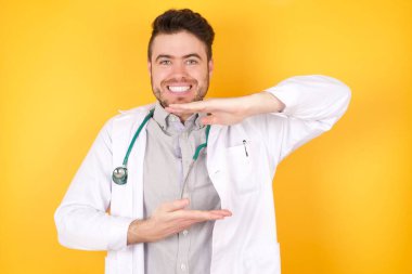 Sarı arka plan üzerinde tıbbi üniforma giyen yakışıklı genç bir doktor. Ellerinde büyük ve büyük boy işareti, ölçü sembolü var. Kameraya bakıp gülümsüyor. Ölçüm konsepti.