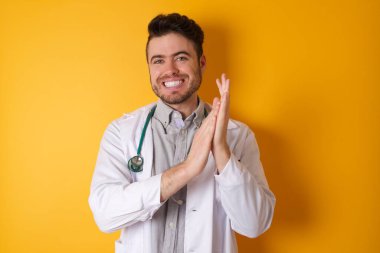 Sarı arka plan üzerinde tıbbi üniforma giyen genç yakışıklı doktor alkışlıyor ve mutlu ve mutlu, gururlu elleri alkışlıyor..