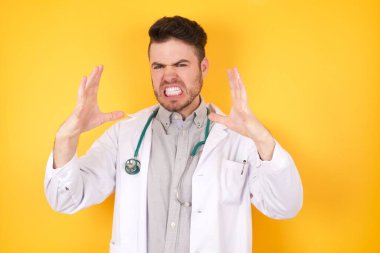 Sarı izole arka planda tıbbi üniforma giyen yakışıklı beyaz bir adam. Öfkeyle bağırıyor, eller boğmaya çalışıyor, deli gibi bağırıyor..