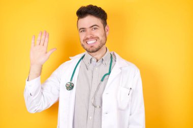 Tıbbi üniforma ve steteskop giyen yakışıklı genç bir doktor, sarı izole edilmiş arka planda. Mutlu ve güler yüzlü el sallayarak, hoş geldin jesti.