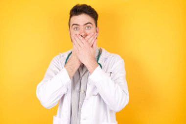 Sarı arka planda tıbbi üniforma giyen yakışıklı beyaz bir doktor yanlışlıkla ağzı ellerle kapatmış. Gizli konsept.