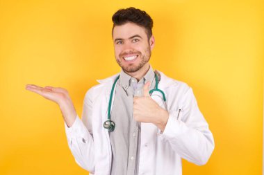 Tıp üniforması ve steteskop giyen yakışıklı beyaz bir adam, avuç içini gösteriyor ve baş parmaklarıyla hareket ediyor, mutlu ve neşeli gülümsüyor..