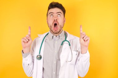 Tıp üniforması giyen yakışıklı beyaz bir doktor şaşırmış ve şaşırmış bir şekilde yukarı bakıp, parmaklarını göstermiş ve kollarını kaldırmış..
