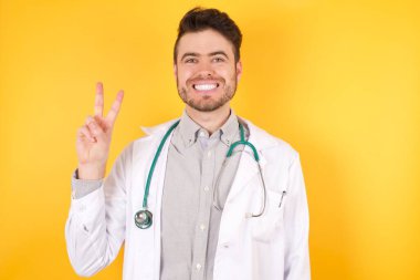 Genç, Avrupalı yakışıklı doktor üniformalı adam izole edilmiş sarı arka plan üzerinde duruyor ve iki numarayı işaret ederken kendinden emin ve mutlu bir şekilde gülümsüyor..