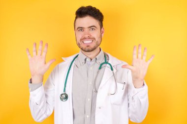 Tıbbi giysiler ve steteskop giyen beyaz arka plan üzerinde duran ve on numaralı parmaklarıyla işaret eden, kendinden emin ve mutlu gülümseyen beyaz saçlı yakışıklı doktor..