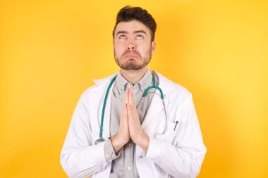 İzole edilmiş sarı arka plan üzerinde tıbbi üniforma giyen genç beyaz doktor yüzünde umut ifadesi olan ve endişeli bir şekilde el ele yalvarıp dua ediyor. Soruyorum.