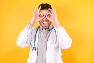 Tıbbi üniforma giyen yakışıklı beyaz bir doktor, izole edilmiş sarı bir geçmişi var. Dürbün gibi güzel hareketler yapıyor. Gözleri parmaklarından bakıyor. Çılgın ifade.