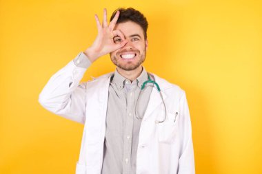 Tıbbi üniforma giyen yakışıklı beyaz bir doktor, izole edilmiş beyaz arka planda duruyor. El hareketleriyle gülümsüyor, gözleriyle mutlu yüzle parmaklarının arasından bakıyor..