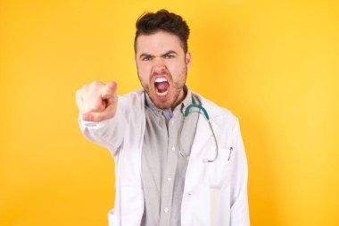 Tıbbi üniforma giyen yakışıklı beyaz bir doktor izole edilmiş sarı arka planda dikiliyor. Kameraya kızgın ve sinirli bir şekilde sizinle kavga etmeye hazır olduğunu gösteriyor..
