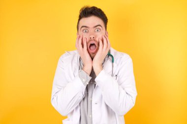 Şaşkın, yakışıklı, beyaz, doktor üniformalı, heyecanlı ve heyecanlı, çenesi düşük, elleri yanaklarında, gözleri fırlamış..