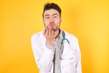 Tıp üniforması giyen yakışıklı, beyaz bir doktor kameraya bakıyor ve havada el ele öpüşerek sevimli ve seksi oluyor. Aşk ifadesi.