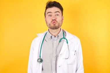 Hoş görünümlü doktor dudaklarını büzüyor, kameraya yeşil gözlü bakıyor, tıbbi üniforma giyiyor, sarı duvarın arkasında izole edilmiş biriyle eğleniyor. İnsan yüz ifadeleri