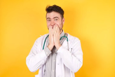 Genç, hayat dolu yakışıklı doktor üniforması ile kıkırdıyor, neşe içinde kıkırdıyor, ağzını kapatıyor, tek başına eğleniyor, beyaz arka plan veya stüdyo duvarına karşı duruyor, doğal kahkahaları var, pozitif hikayeler duyuyor ya da komik anekdotlar dinliyor.