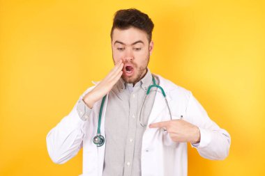 Şok olmuş genç, yakışıklı doktor üniformalı adam şaşırtıcı bir şekilde yere bakıyor işaret parmağıyla boş bir alanda stüdyonun arka planında poz veriyor.