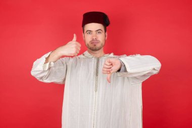 Geleneksel Türk şapkası takan yakışıklı genç adam başparmağını kaldırıyor ve başparmağını eğiyor, zor seçim konsepti.  