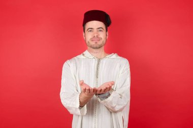 Geleneksel Türk şapkası takan yakışıklı genç adam elinde avuç içi açık bir şeyle kameraya sunuyor..