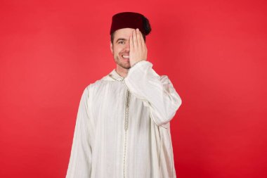 Fes takan yakışıklı genç bir adam bir gözünü eliyle kapatıyor, yüzünde kendine güvenen bir gülümseme ve şaşırtıcı bir duygu var..