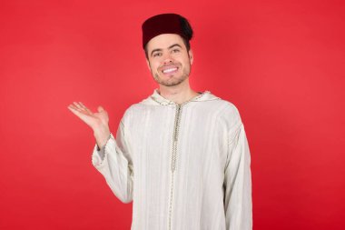 Fes takan yakışıklı genç adam izole edilmiş kırmızı arka planda gülümsüyor ve avucunun içi gibi kameraya bakıyor..
