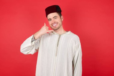 Fes takan yakışıklı genç adam telefonla konuşuyor gibi el ve parmaklarıyla telefon hareketi yapıyor. İletişim kavramları.