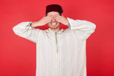 Fes takan yakışıklı genç adam elleri ile gözleri kapatıyor neşeli ve komik gülümsüyor. Kör kavram.