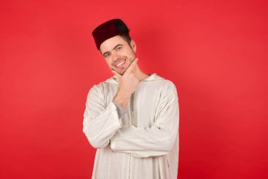 Fes takan yakışıklı genç adam kameraya kendinden emin bakıyor kollarını kavuşturmuş ve çenesini kaldırmış gülümsüyor. Olumlu düşünüyorum..