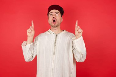 Fes takan yakışıklı genç adam şaşırmış ve şaşırmış yukarı bakıyor ve parmaklarıyla işaret ediyor ve kollarını kaldırıyor..