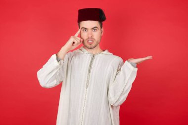 Geleneksel Türk şapkası takan yakışıklı genç adam avucunun içi açık, fotokopi alanını gösteren ve parmağıyla alnını gösteren fes şapkası takıyor. Bunu bir düşün..