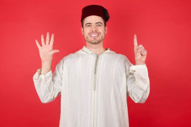 Geleneksel Türk şapkası giyen yakışıklı genç adam altı numaralı parmaklarıyla kendini beğenmiş ve mutlu gülümserken kendini gösteriyor..