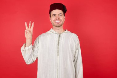 Geleneksel Türk şapkası giyen yakışıklı genç adam üç numaralı parmaklarıyla kendini beğenmiş ve mutlu gülümserken görülüyor..