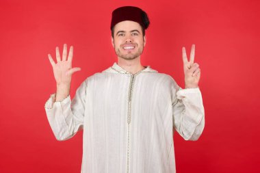 Geleneksel Türk şapkası giyen yakışıklı genç adam yedinci parmaklarıyla kendini beğenmiş ve mutlu gülümserken görülüyor..