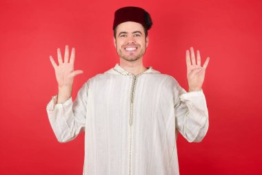 Geleneksel Türk şapkası giyen yakışıklı genç adam dokuz numaralı parmaklarıyla kendini beğenmiş ve mutlu gülümserken görülüyor..