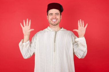 Geleneksel Türk şapkası giyen yakışıklı genç adam on numaralı parmaklarıyla kendini beğenmiş ve mutlu gülümserken kendini gösterdi..