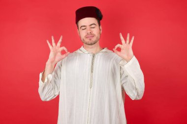 Geleneksel Türk şapkası takan yakışıklı genç adam gözleri kapalı gülümsüyor parmaklarıyla meditasyon hareketi yapıyor. Yoga konsepti.