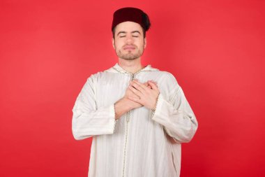Geleneksel Türk şapkası takan yakışıklı genç adam elleri göğsünde ve gözleri kapalı gülümsüyor. Sağlık kavramı.