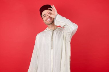 Geleneksel Türk şapkası takan yakışıklı genç adam el hareketleriyle gülüyor ve mutlu yüzüyle parmaklarının arasından bakıyor..