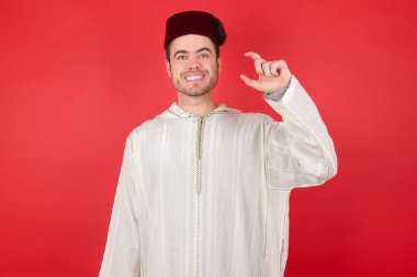 Geleneksel Türk şapkası takan yakışıklı genç bir adam gülümseyerek ve el hareketleriyle kendinden emin bir şekilde hareket ediyor, parmaklarıyla küçük boy işareti yapıyor ve kameraya bakıyor. Ölçü kavramı