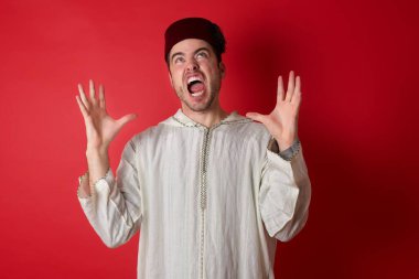 Geleneksel Türk şapkası giyen yakışıklı genç adam deli gibi bağırıyor ve agresif bir ifadeyle bağırıyor ve kollarını kaldırıyor. Hayal kırıklığı konsepti.
