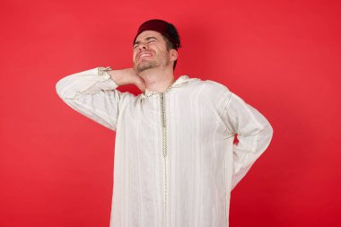 Geleneksel Türk şapkası takan yakışıklı genç adam boyun ağrısı çekiyor, boynuna elle dokunuyor, kas ağrısı çekiyor.