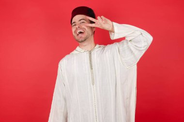 Geleneksel Türk şapkası takan yakışıklı genç bir adam barış sembolü yapıyor. Yüzünde parmaklar, gülümseyerek zaferi gösteriyor.