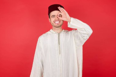 Geleneksel Türk şapkası takan yakışıklı genç adam el hareketleriyle gülüyor ve mutlu yüzüyle parmaklarının arasından bakıyor..