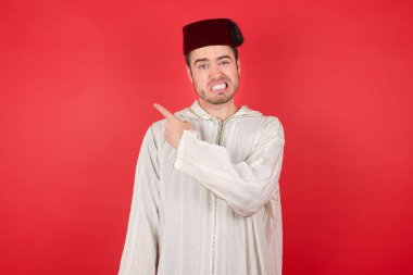 Geleneksel Türk şapkası takan yakışıklı genç adam endişeli ve endişeli bir kenara işaret ediyor işaret parmağı, endişe ve sürpriz konsept.