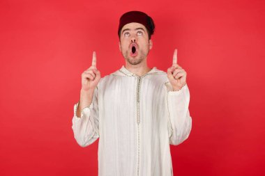 Geleneksel Türk şapkası giyen yakışıklı genç adam şaşırmış ve şaşırmış bir şekilde yukarı baktı ve parmaklarıyla işaret etti ve kollarını kaldırdı..