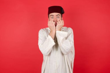 Geleneksel Türk şapkası giyen yakışıklı genç adam Fez ellerini ağzının yakınında tutuyor, korkmuş ve korkmuş hissediyor, kırmızı gömlek giyen, Afro saç stiline sahip bir fobisi var. Şok ve korku konsepti.