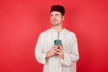 Geleneksel Türk şapkası takan yakışıklı genç adam kırmızı arka planda akıllı telefon kullanıyor.