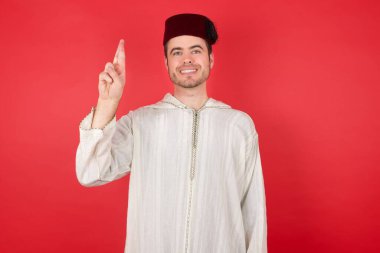 Kırmızı stüdyo arka planında poz veren, fes takan yakışıklı genç bir adam portresi.