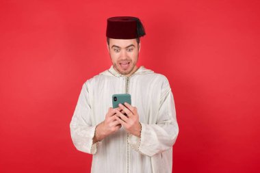 Geleneksel Türk şapkası takan yakışıklı genç adam kırmızı arka planda akıllı telefon kullanıyor.